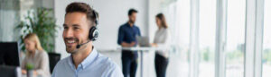 Smiling customer service agent wearing a headset in the foreground of a bright office.